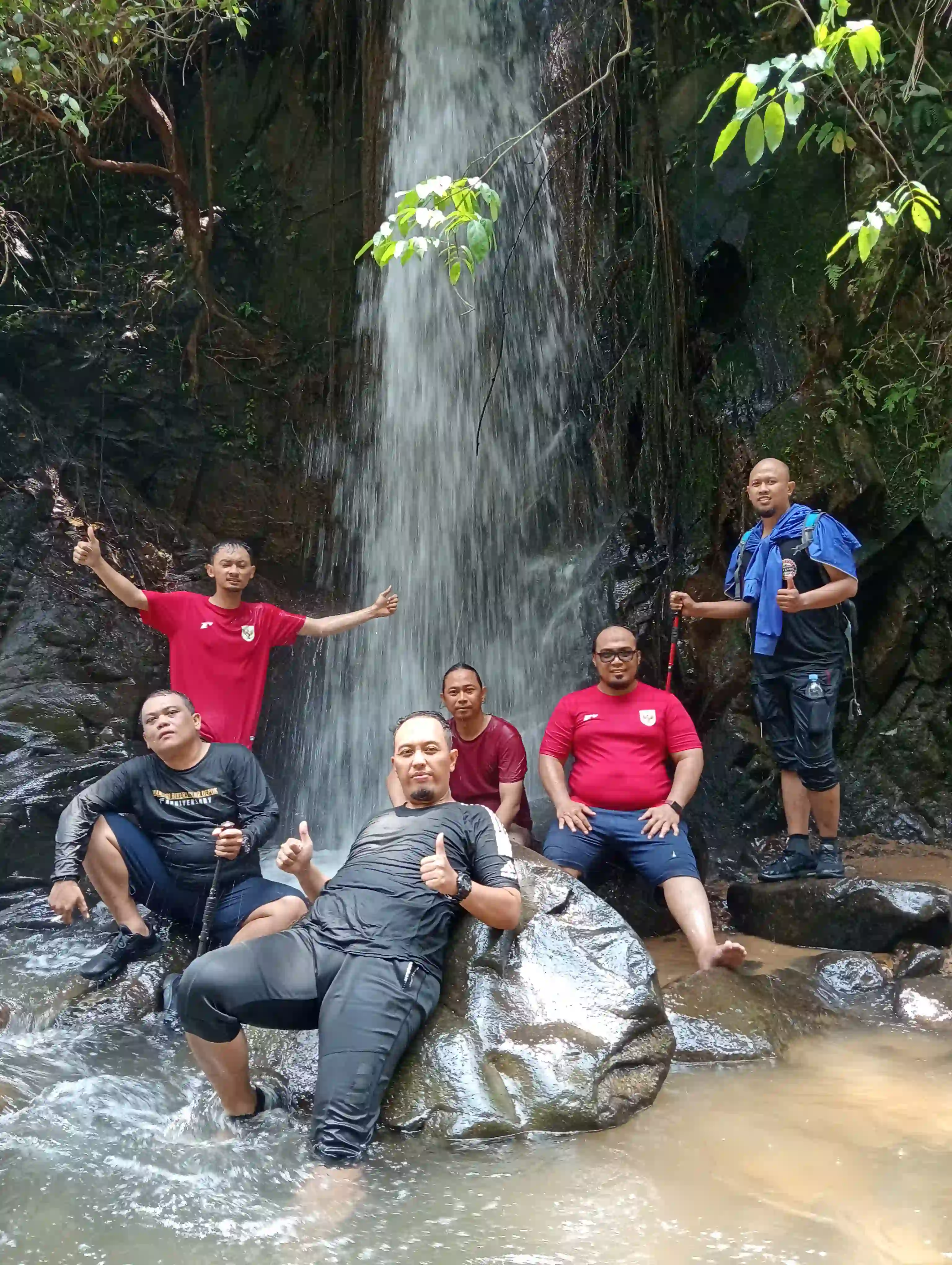 Lima peserta pria bersantai dan berpose di atas bebatuan sungai di bawah air terjun.