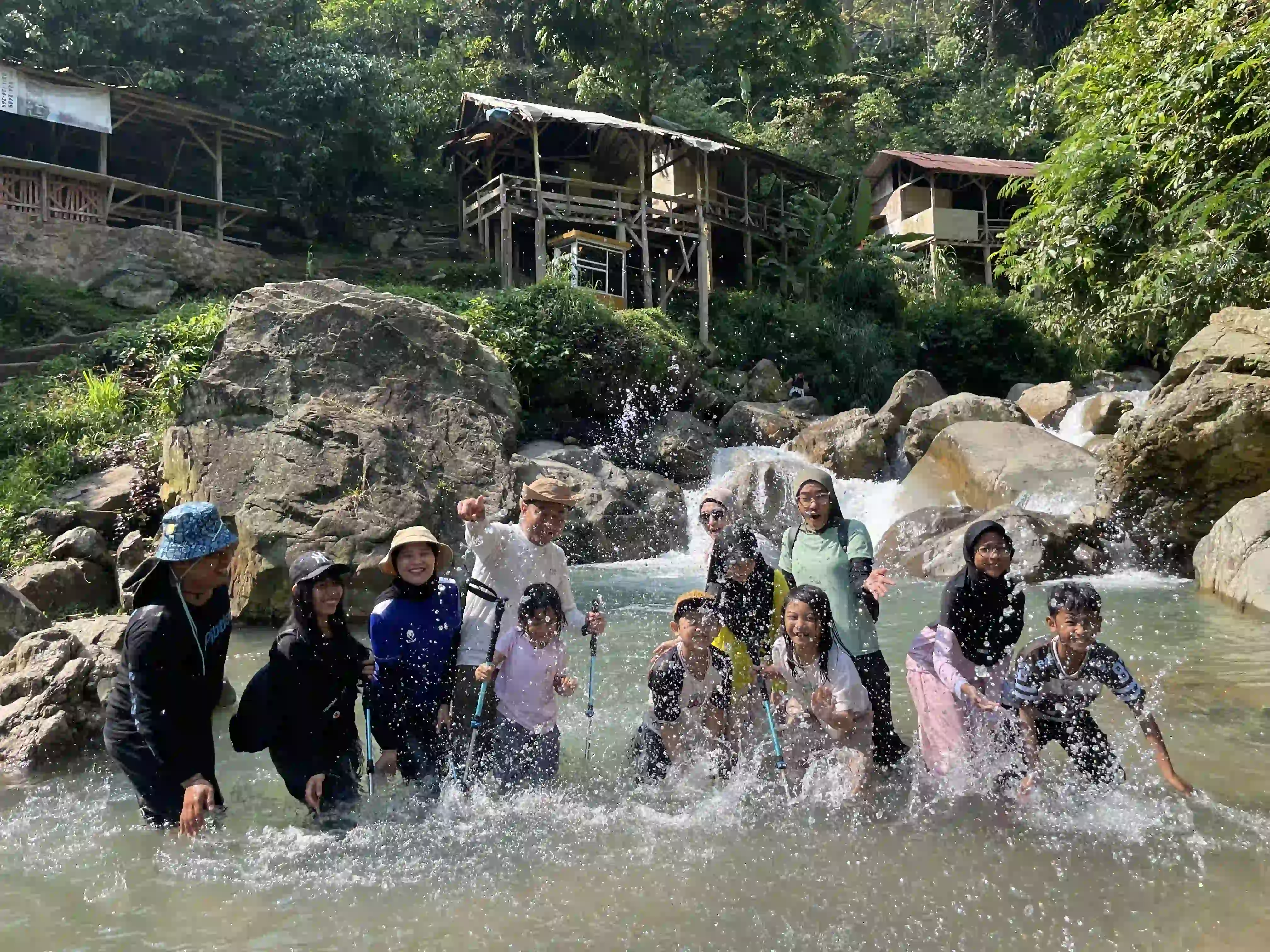 Peserta trekking berfoto bersama sambil mencipratkan air di sungai dengan latar bangunan kayu tradisional.