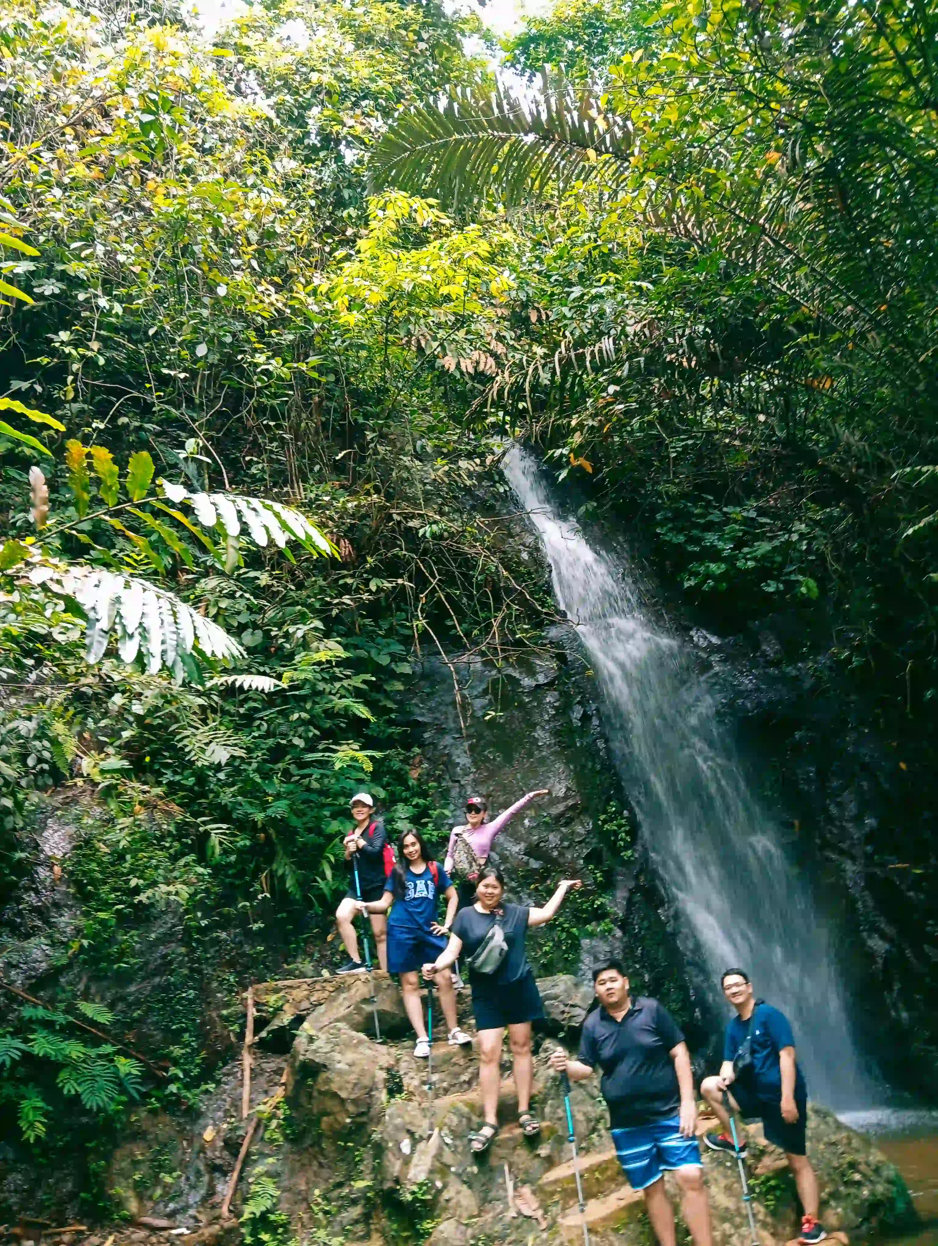 Sekelompok peserta berpose di atas bebatuan besar di kaki air terjun yang dikelilingi vegetasi hijau.