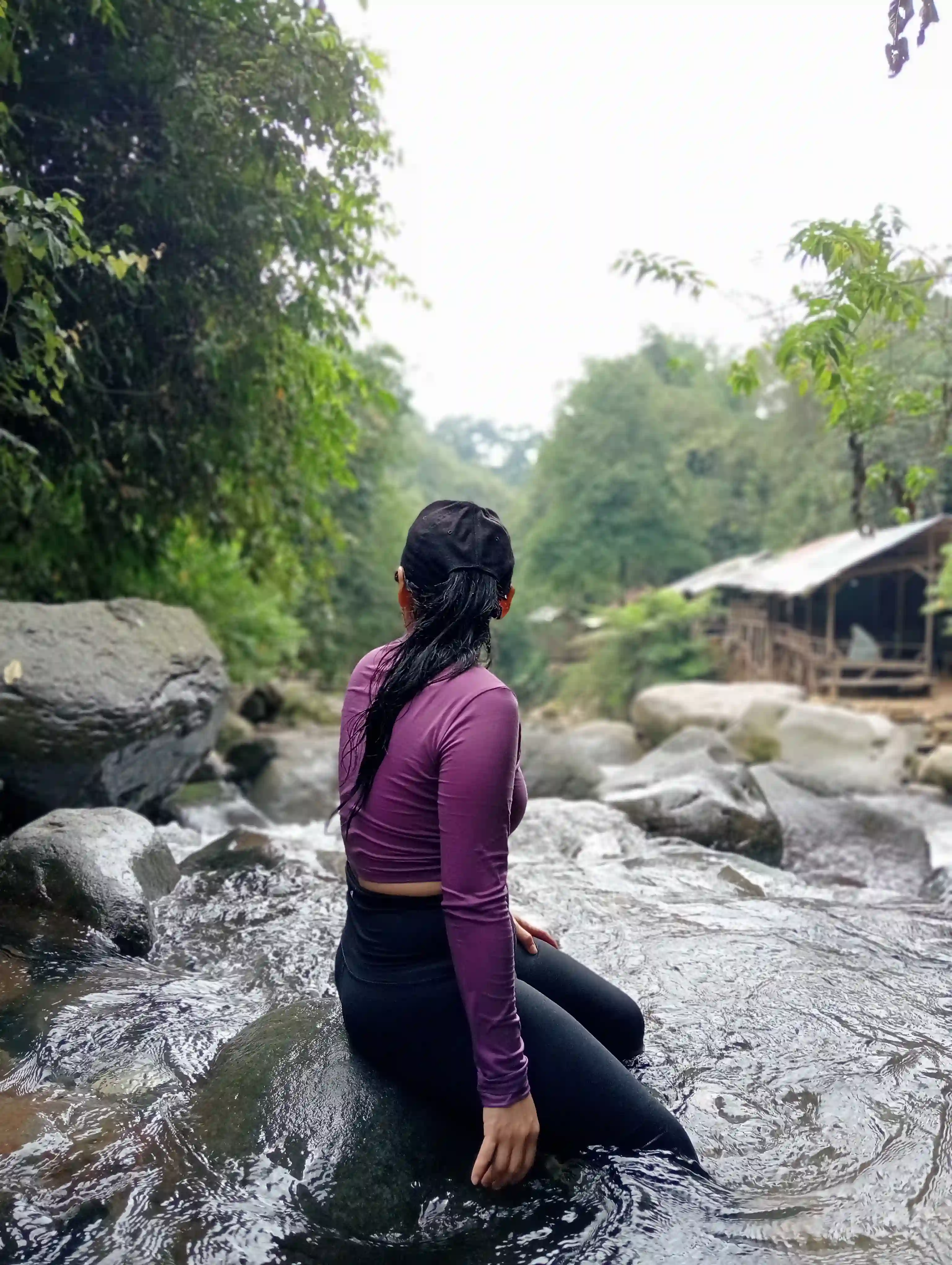 Peserta wanita duduk menghadap aliran sungai dengan latar belakang pondok kayu tradisional di kejauhan.